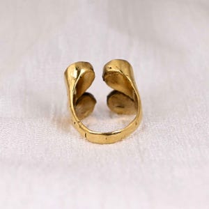 May include: A gold-colored open ring with a unique design. The ring features two curved, leaf-like shapes that meet at the top, creating an open space. The ring is set against a white background, highlighting its metallic sheen.