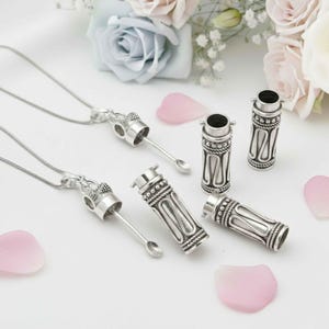 Puede incluir: Collares con colgantes de metal plateado y recipientes cilíndricos. Los colgantes tienen un diseño en forma de cuchara. Los recipientes tienen patrones decorativos y se muestran con pétalos de flores rosas y un arreglo floral.