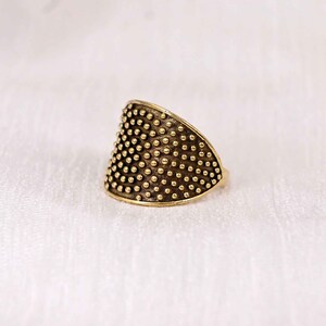 Può includere: Un anello color oro con una superficie strutturata. L'anello presenta un design triangolare ricoperto da piccoli punti in rilievo. L'anello è posto su uno sfondo bianco, evidenziando i suoi dettagli intricati. Questo gioiello unico è adatto all'uso quotidiano.