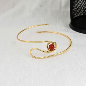 May include: Gold-toned cuff bracelet featuring a central, round, reddish-orange stone. The bracelet has a minimalist design with a curved, open shape. The stone is held in place by a delicate wire wrap, adding a touch of elegance to the jewelry.