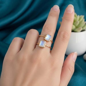 May include: Two gold rings, each featuring a rectangular, iridescent gemstone. The rings are displayed on a hand, with a teal background and a small potted plant visible in the background. The gemstones have a blue and white color.