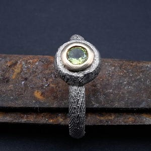 May include: A textured silver ring with a gold bezel setting. The ring features a round, light green gemstone. The band has a detailed, woven pattern. The ring is displayed on a rusty, textured surface.