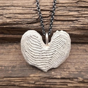 May include: A silver-colored pendant in the shape of a heart, suspended from a matching chain. The heart pendant has a textured surface with horizontal lines, giving it a unique, organic appearance. The chain is a dark silver color, and the pendant hangs against a wooden background.