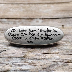 May include: A silver-coloured, oval-shaped stone with black handwritten text. The text reads, "Du bist kein Tropfen im Ozean. Du bist ein gesamter Ozean in einem Tropfen." The stone rests on a weathered wooden surface.