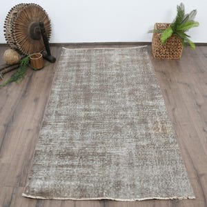 May include: A rectangular, distressed rug in shades of grey and beige. The rug has a faded, vintage appearance with a textured surface. The rug is placed on a wooden floor, with decorative items in the background.