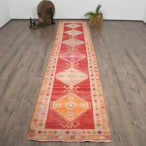 Peut inclure: Un long tapis rectangulaire rouge avec des motifs géométriques orange, rose et crème. Le tapis présente un motif central en losange et une bordure avec des motifs répétitifs. Ce tapis vintage convient aux couloirs ou aux entrées.