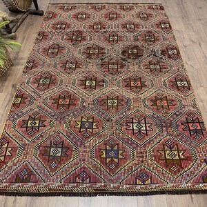 May include: A rectangular rug with a geometric pattern in shades of red, brown, purple, yellow, and blue. The rug features a repeating diamond and star design, with a fringed edge. The rug is placed on a wooden floor.