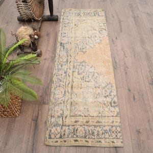 Puede incluir: Una alfombra rectangular de estilo vintage con un aspecto descolorido y desgastado. La alfombra presenta un campo central beige con intrincados patrones florales y geométricos en tonos azul y crema apagados. La alfombra está colocada sobre un suelo de madera.