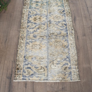 May include: A rectangular, distressed vintage rug with a floral pattern in shades of blue, beige, and brown. The rug has a faded appearance, suggesting age and use. The rug is placed on a wooden floor.