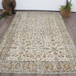 May include: A rectangular, cream-colored rug with an intricate floral pattern. The rug features a border with blue and gold accents. The rug is placed on a dark wood floor, with a basket and decorative items in the background.