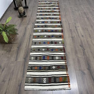 Könnte beinhalten: Ein langer, rechteckiger Läuferteppich mit Streifenmuster. Der Teppich hat abwechselnde Bänder in Weiß, Grau und Schwarz mit farbenfrohen geometrischen Akzenten in Orange, Blau und Grün. Der Teppich liegt auf einem Holzboden.