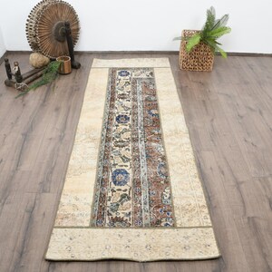 May include: A rectangular beige rug with a central patterned design in shades of blue, brown, and cream. The rug features floral and geometric motifs, with a border of a lighter beige color. The rug is placed on a wooden floor.