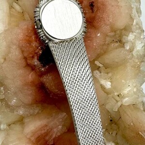 Puede incluir: Un reloj plateado con una correa de malla y una esfera redonda plateada. El reloj est&aacute; sobre una pieza de roca blanca y rosa.