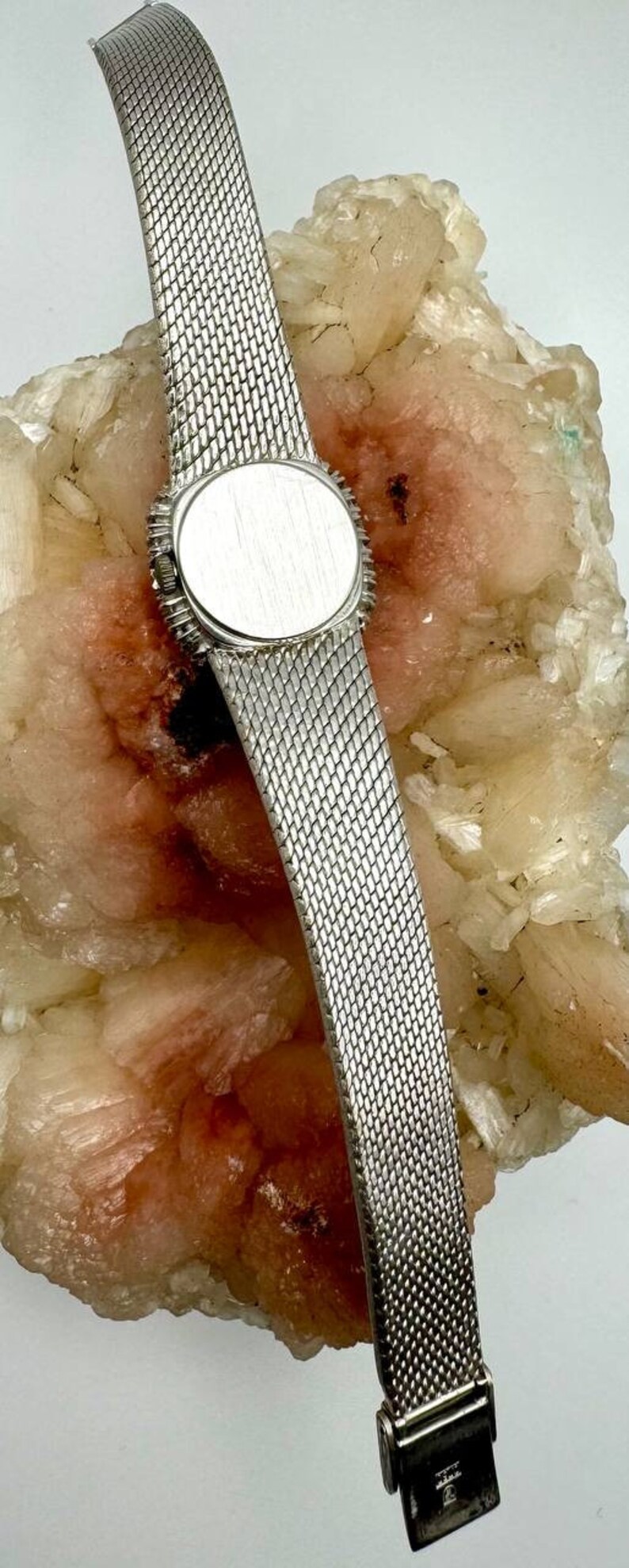 Puede incluir: Un reloj plateado con una correa de malla y una esfera redonda plateada. El reloj est&aacute; sobre una pieza de roca blanca y rosa.