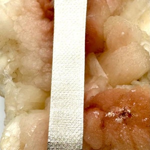 Puede incluir: Un reloj plateado con una esfera cuadrada y un bisel de diamantes. El reloj tiene una correa de cuero blanca con un patr&oacute;n texturizado. El reloj est&aacute; sentado sobre una roca rosa y blanca.