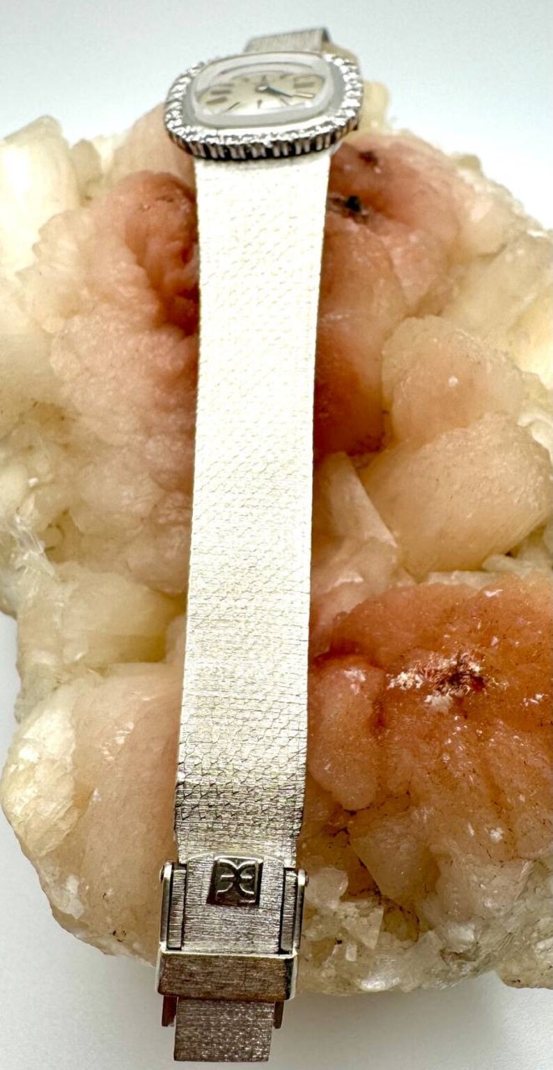 Puede incluir: Un reloj plateado con una esfera cuadrada y un bisel de diamantes. El reloj tiene una correa de cuero blanca con un patr&oacute;n texturizado. El reloj est&aacute; sentado sobre una roca rosa y blanca.