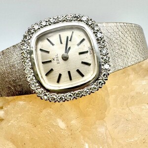 Puede incluir: Un reloj de pulsera plateado con una esfera cuadrada rodeada de diamantes. La esfera del reloj tiene agujas negras y marcas de n&uacute;meros romanos. La correa del reloj es plateada y texturizada. La esfera del reloj dice "Ebel" y "Swiss".