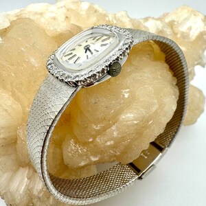 Puede incluir: Un reloj plateado con una esfera cuadrada y un bisel de diamantes. El reloj tiene una esfera blanca con marcadores de hora en n&uacute;meros romanos negros y una correa de malla plateada.