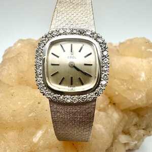 Puede incluir: Un reloj de pulsera Ebel plateado con una esfera cuadrada y un bisel engastado con diamantes. El reloj tiene una esfera blanca con agujas e &iacute;ndices negros. El reloj est&aacute; en una pulsera de metal plateado.