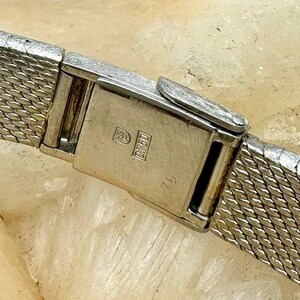 Puede incluir: Primer plano de una correa de reloj plateada con un cierre que tiene la inscripci&oacute;n "0750" y un s&iacute;mbolo.