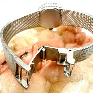 Puede incluir: Un reloj plateado con una correa de malla y un cierre de hebilla. El reloj tiene una esfera redonda y un bisel plateado.