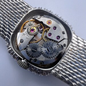 Puede incluir: Un reloj plateado con una correa de malla plateada. La parte posterior del reloj est&aacute; abierta, revelando el funcionamiento interno del reloj. La esfera del reloj tiene el texto "Ebel Fab. Suisse" impreso en ella.