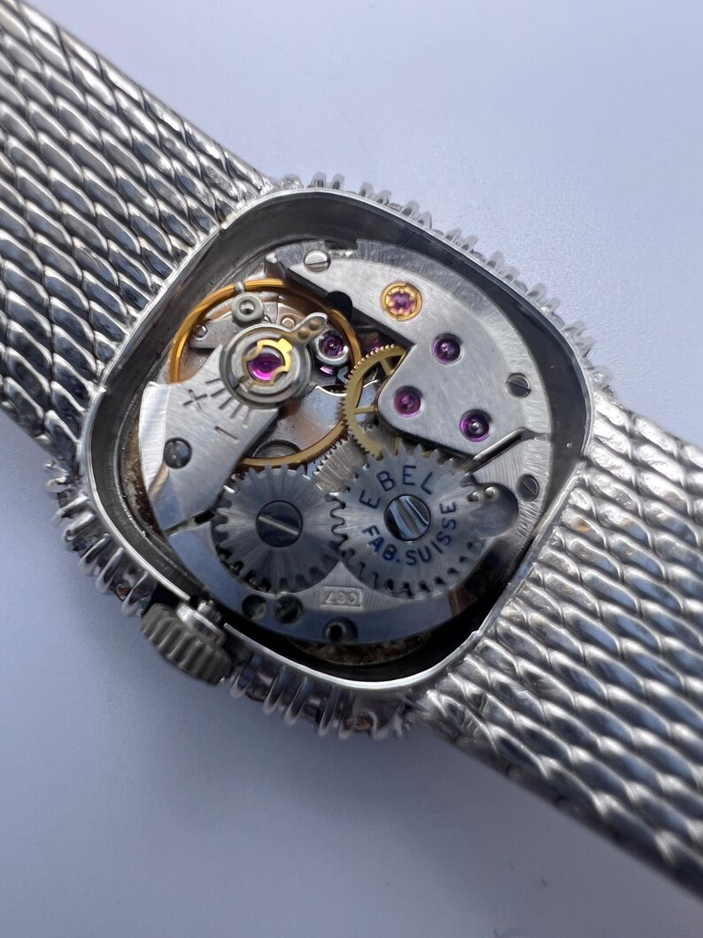 Puede incluir: Un reloj plateado con una correa de malla plateada. La parte posterior del reloj est&aacute; abierta, revelando el funcionamiento interno del reloj. La esfera del reloj tiene el texto "Ebel Fab. Suisse" impreso en ella.