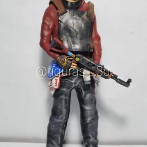May include: A hand-painted action figure of a person wearing a red hooded sweatshirt, a metal chest plate, a black mask, and blue jeans. The figure is holding a black rifle with a yellow smiley face on the side. The figure is standing on a white surface. The text "@figuras_3d" is visible in the lower left corner of the image.