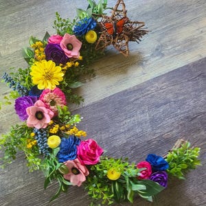 May include: A crescent-shaped floral wreath with a variety of colorful flowers, including pink, yellow, blue, and purple blooms. A woven star with an orange butterfly sits at the top. Green foliage accents the flowers.