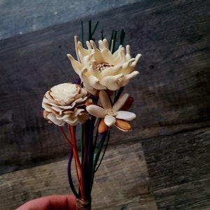 Peut inclure: Un bouquet de fleurs séchées aux pétales blancs et bruns, lié avec de la ficelle et tenu par une main. Les tiges sont enveloppées de ficelle colorée.