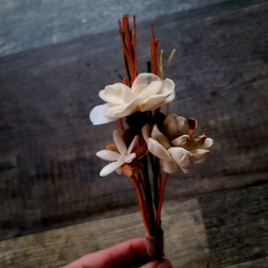 Puede incluir: Un ramo de flores blancas secas con tallos marrones atados con cuerda. Las flores están dispuestas en una mano.