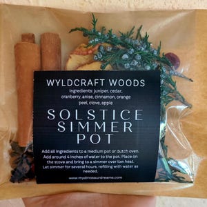 May include: A clear cellophane bag containing ingredients for a Solstice Simmer Pot. The bag includes cinnamon sticks, dried apple slices, juniper, cedar, and a black label with white text that reads "WYLDCRAFT WOODS SOLSTICE SIMMER POT".