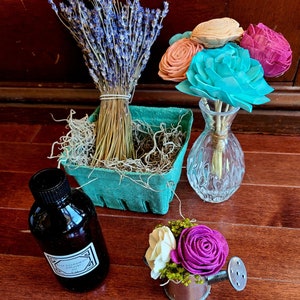 Puede incluir: Un ramo de flores de lavanda secas en una cesta verde, una pequeña botella de aceite difusor de sándalo y un ramo de flores artificiales de colores en un jarrón de vidrio transparente. Las flores están dispuestas en una pequeña regadera.