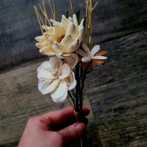 以下が含まれることがあります： 白とクリーム色の花と茶色のアクセントが付いたドライフラワーのブーケ。ブーケは紐で結ばれており、緑色の茎があります。