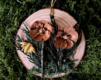 May include: A circular wooden ornament with a natural wood grain, decorated with two brown pinecone flowers and a gold leaf. Green foliage and moss surround the flowers. A gold string is attached for hanging. The background is a green textured surface.