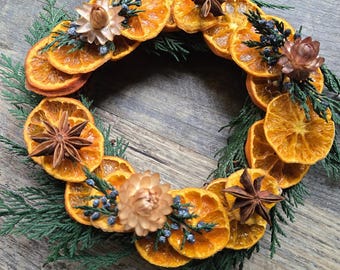 May include: A circular wreath featuring dried orange slices, star anise, and evergreen sprigs. The wreath is adorned with small, dried flowers and blue berries. The arrangement is set against a wooden background, creating a rustic and natural aesthetic.