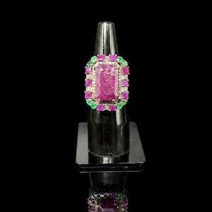 May include: A rectangular ring featuring a large central ruby surrounded by a border of small square-cut rubies and round green emeralds. The setting is metallic, with the stones arranged in a structured, multi-layered geometric pattern.