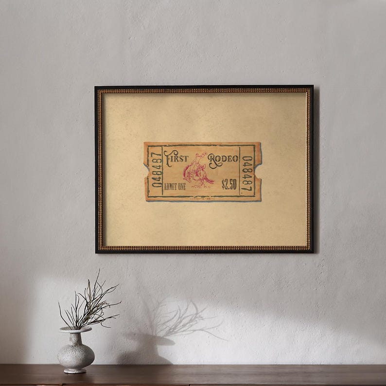 May include: Framed vintage rodeo ticket with the text "First Rodeo" and a graphic of a rodeo rider. The ticket is tan with red and black text, framed in a black and gold ornate frame. The artwork is displayed on a light beige background.