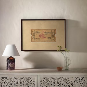 May include: A framed vintage rodeo ticket with the text "First Rodeo" and a graphic of a bull rider. A small table lamp with a white shade and a decorative base sits to the left. A vase with flowers and a small bowl are on the table.