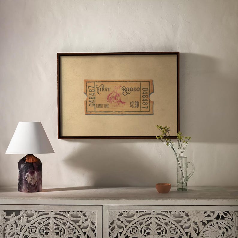 May include: A framed vintage rodeo ticket with the text "First Rodeo" and a graphic of a bull rider. A small table lamp with a white shade and a decorative base sits to the left. A vase with flowers and a small bowl are on the table.