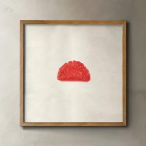 May include: A framed print featuring a single, vibrant red gummy candy in a half-circle shape. The candy is centered on a white background, encased in a natural wood frame.