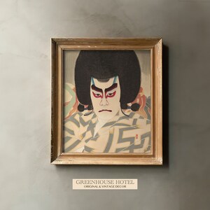 Puede incluir: Una pintura enmarcada de un hombre con una afro negra, vistiendo una t&uacute;nica estampada y un maquillaje dram&aacute;tico. La pintura est&aacute; en un marco de madera con un acabado marr&oacute;n claro. El texto "GREENHOUSE HOTEL ORIGINAL & VINTAGE DECOR" est&aacute; debajo de la pintura.