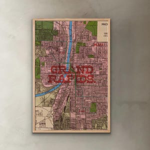 May include: A framed vintage map of Grand Rapids, Michigan, in a light wood frame. The map features a pink and green colour scheme with a blue river running through the centre. The city name is prominently displayed in red block letters.
