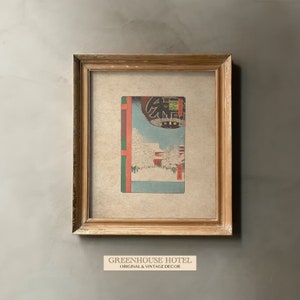 Puede incluir: Una impresi&oacute;n japonesa vintage enmarcada que representa una escena nevada con una puerta torii roja y una linterna. La impresi&oacute;n est&aacute; en un marco de madera claro y tiene un passe-partout de color crema.