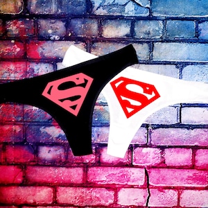 Könnte beinhalten: Zwei Paar schwarze und weiße Unterwäsche mit einem Superman-Logo. Ein Paar hat ein pinkfarbenes Superman-Logo und das andere ein rotes Superman-Logo.
