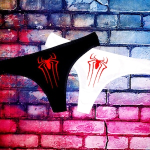 Könnte beinhalten: Schwarzer und weißer String-Tanga mit roter Spinnen-Grafik. Die Unterwäsche wird vor einer Backsteinmauer mit einem farbenfrohen Graffiti-Design präsentiert. Die Spinnengrafik ist ein stilisiertes Design.