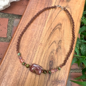May include: A handmade necklace featuring a brown macrame cord with a square, reddish-brown stone pendant. The necklace is adorned with green, pink, and brown beads, creating a natural and earthy aesthetic. The clasp is silver-toned.