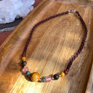 May include: A handmade necklace featuring a dark brown woven cord and a variety of colorful beads. The beads include shades of yellow, red, green, pink, and a large, round, orange-brown bead. The necklace is displayed on a wooden tray.