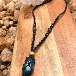 May include: A handmade macrame necklace featuring a teal stone pendant. The necklace has a black cord with decorative beads and a wooden bead. The pendant is encased in a black macrame net. A clear crystal is in the background.