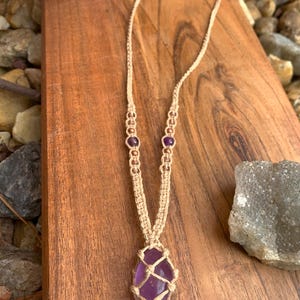 May include: A macrame necklace featuring a large, polished amethyst gemstone. The necklace has a beige cord with small, round beads and two amethyst beads near the top. The gemstone is a deep purple color and is encased in a macrame net.